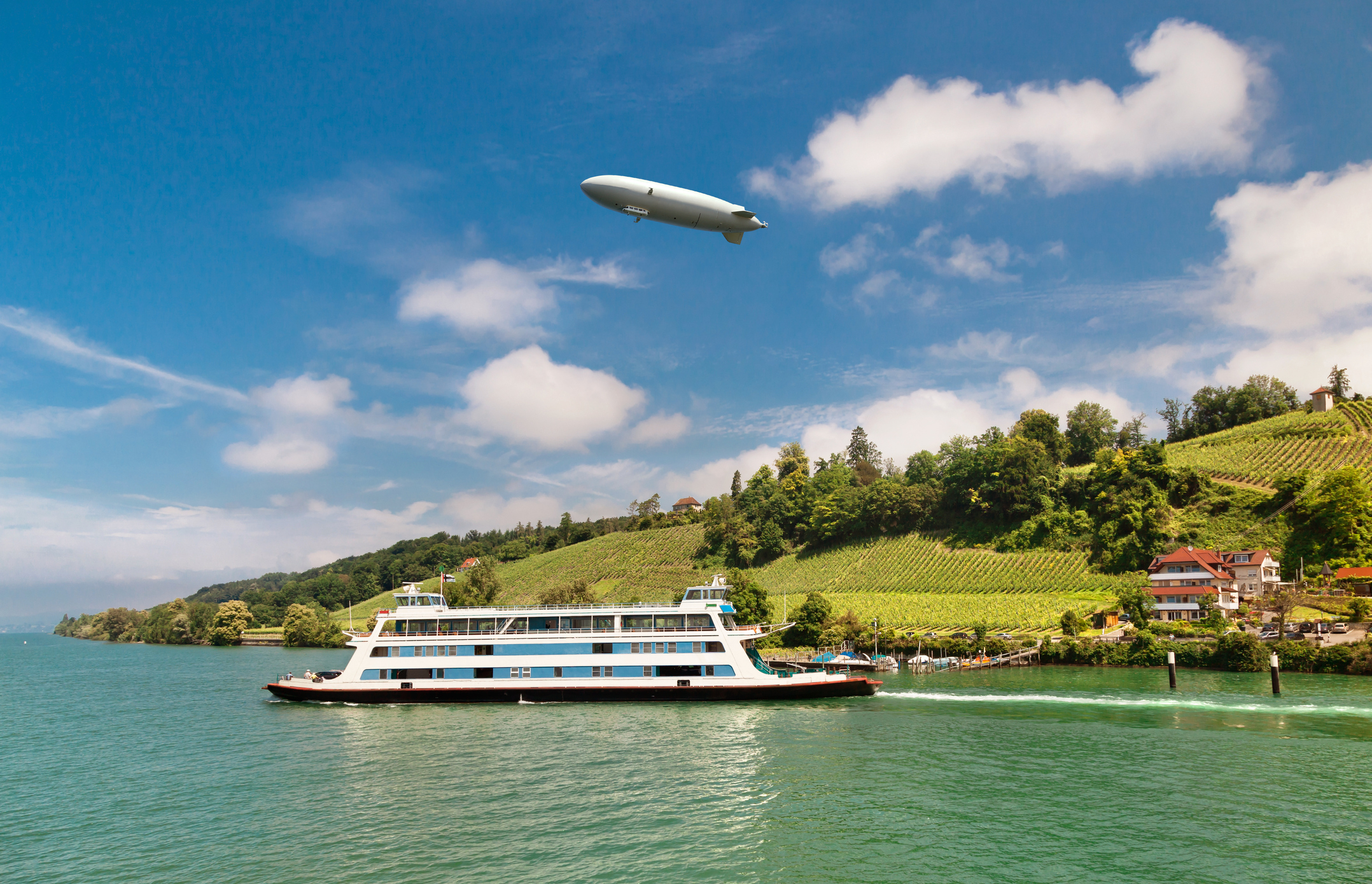 En Zeppelin svæver over et flodlandskab med vinmarker og en færge.