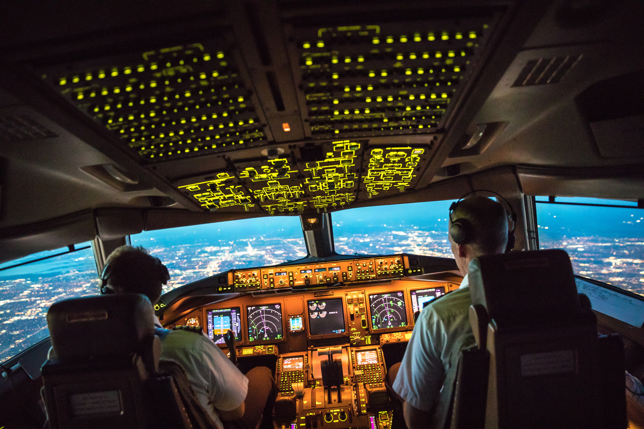 Et oplyst flycockpit med to piloter, der nærmer sig en by om aftenen.