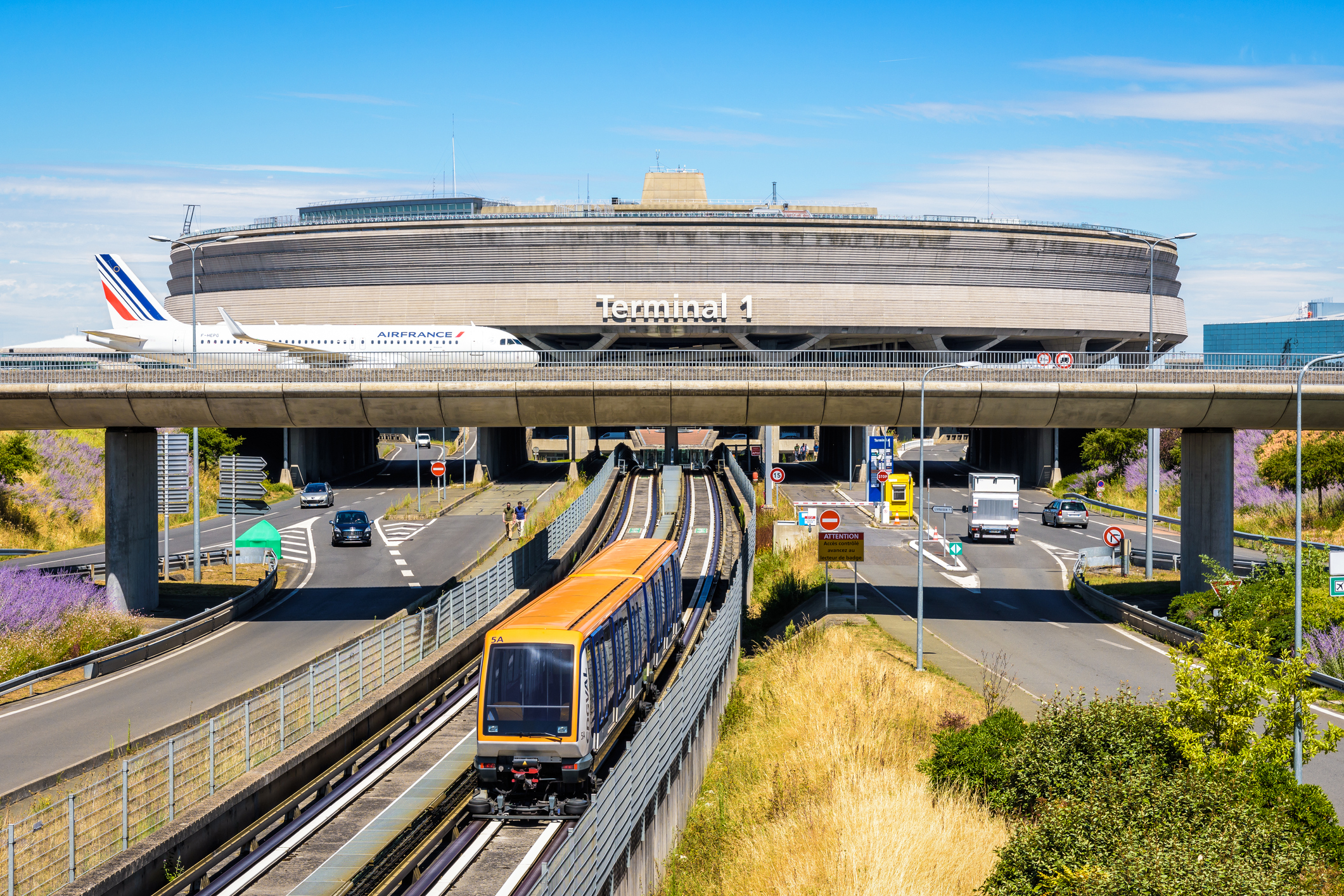 Et billede af Terminal 1 i Charles de Gaulle Airport, en sporvogn kan ses i forgrunden, et Air France-fly krydser en motorvej på en bro i baggrunden.