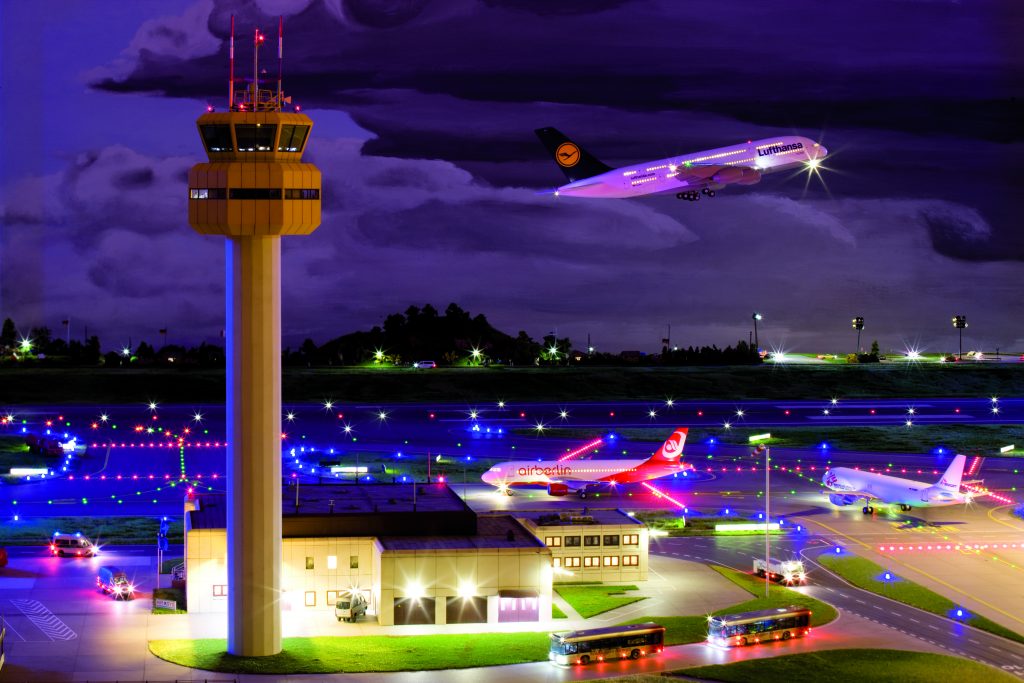 Tårnet i Knuffingen Lufthavn om natten med et Lufthansa-fly, der letter i baggrunden, og to andre fly, der er på vej til startbanen.