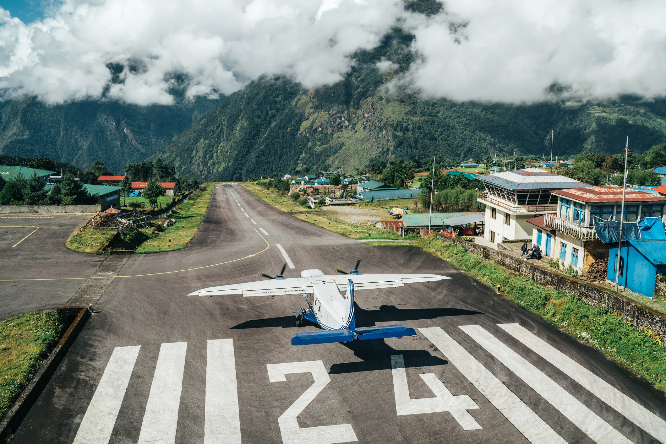 Startbanen i Lukla med solskin, men også nogle lavt hængende skyer, et propel-fly er lige begyndt at lette.