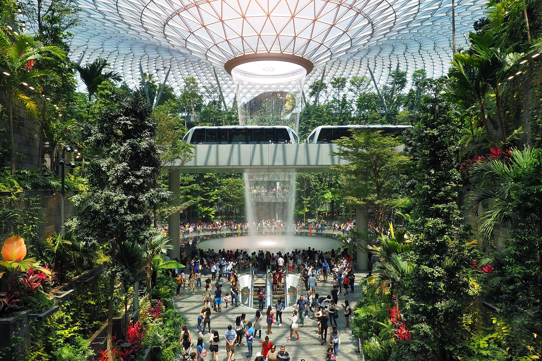 Et panoramabillede inde fra Jewel Changi, der viser Rain Vortex-vandfaldet, tropiske planter og mange rejsende.