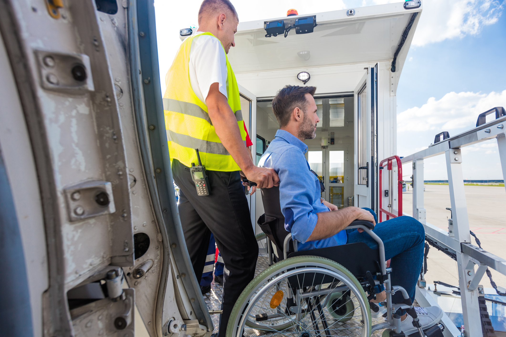 En mand i kørestol bliver skubbet af en ansat i et flyselskab; man kan se flydøren i forgrunden og landgangen i baggrunden.
