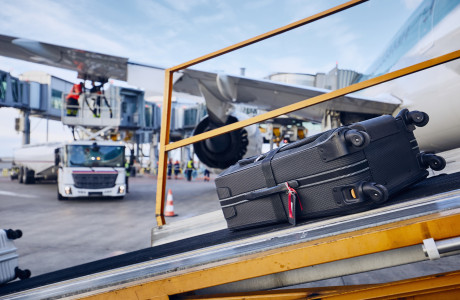 En kuffert læsses på et fly med et bagagebånd, i baggrunden er flyet ved at blive tanket op.